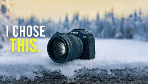 Canon EOS camera with lens on snow-covered ground, evergreen forest in background.