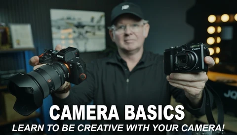 Photography instructor holding a mirrorless camera and compact camera while demonstrating equipment in a studio setting.