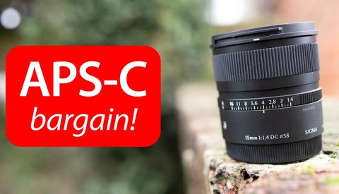 Sigma 15mm f/1.4 DC HSM Art lens positioned on a textured surface with a red banner overlay promoting an APS-C bargain.