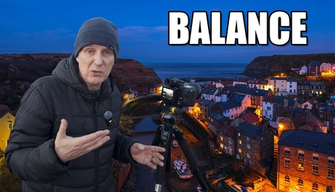 Photographer gesturing while holding a camera on a tripod overlooking a coastal town at dusk.