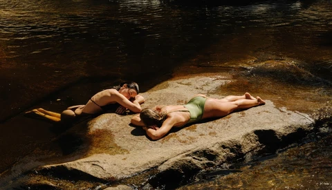 Two people relaxing on a large rock in shallow stream water surrounded by darker water.