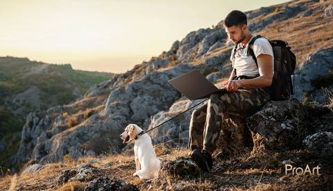 Man sitting on a rocky outcrop working on a laptop with a small dog beside him during golden hour.