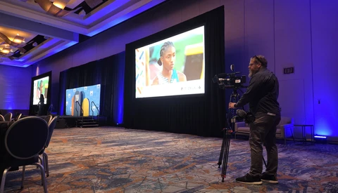 Videographer operating a camera on tripod in a professional event space with blue accent lighting and large projection screens.