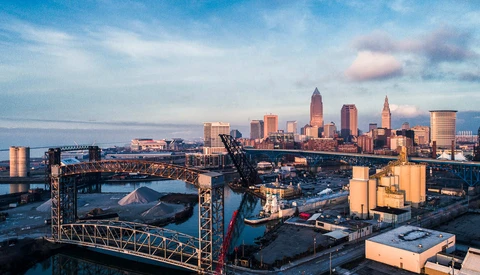 Industrial waterfront at golden hour with city skyline and construction cranes in background.