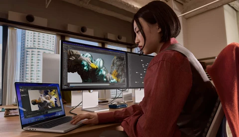 Video editor working on color grading with dual monitors in a bright studio workspace.