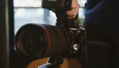 Canon C100 Mark II cinema camera with red-ringed lens held by a photographer indoors.