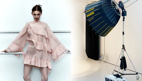 Model wearing layered pink ruffled dress positioned against white wall beside professional lighting setup.