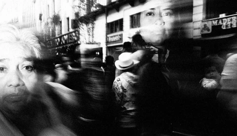 Black and white photograph with intentional motion blur showing multiple faces and figures in an urban street scene.