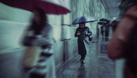 Person with umbrella walking through wet cobblestone alley with blurred figures and buildings