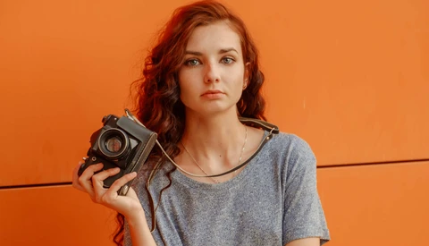 Woman with red wavy hair holding a vintage film camera against an orange background.