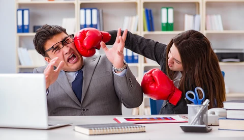 Two business professionals in office attire engage in a playful boxing match at a desk.