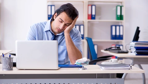 Man in blue shirt leaning on desk, looking tired while working at laptop in office.