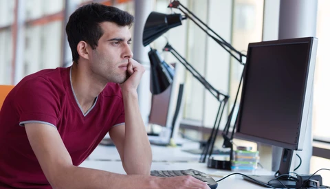 Man in maroon shirt sitting at desk with monitor, appearing thoughtful while working.