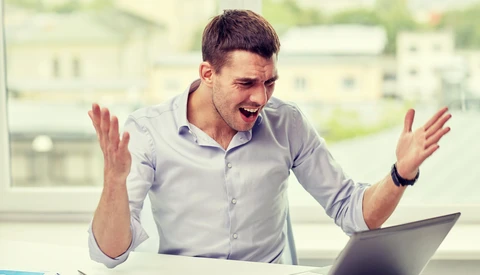 Man at desk gesturing expressively while looking at laptop screen.