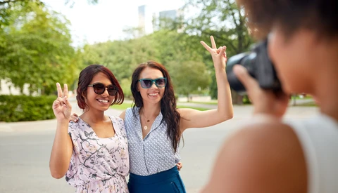 Two women making peace signs while posing for a photo held by a person in the foreground.