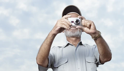Older man with gray beard and cap holding vintage rangefinder camera up to his face against cloudy sky.