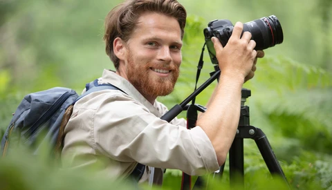 Male photographer with beard operating camera on tripod while smiling at viewer in outdoor forest setting.