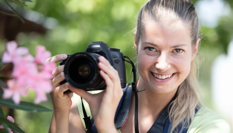 Young female photographer smiling while holding a DSLR camera with a telephoto lens in an outdoor garden setting.
