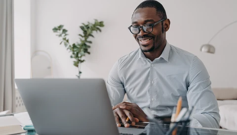 Man in light blue shirt and glasses working at laptop at desk with plants and natural light.