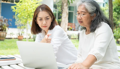 Two women in white shirts reviewing laptop screen outdoors in a garden setting.
