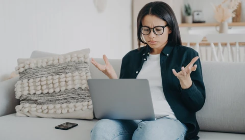 Woman with glasses looking frustrated at laptop screen while sitting on couch.