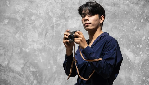 Photographer in dark blue jacket holding a compact camera, posed against a textured gray concrete wall.
