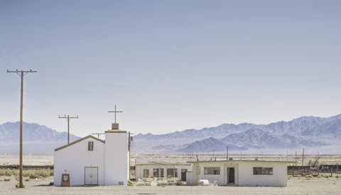 White church and buildings in an arid desert landscape with mountains in the distance.