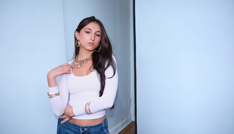 Woman in white cropped long-sleeve top and blue jeans posing against a white wall with geometric shadow.