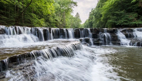 Cascading tiered waterfall flowing through a forested gorge with lush green canopy.