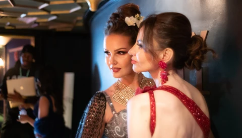 Two women with styled hair and makeup at an indoor event, one wearing a beaded dress and the other in a red ribbon detail dress, backlit by blue lighting.