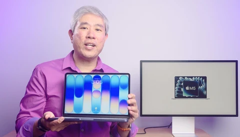 Man in magenta shirt displaying laptop with blue gradient wallpaper beside monitor showing M5 chip graphic.