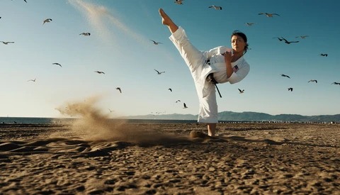 Martial artist in white gi performing a high kick on a sandy beach with birds flying overhead.