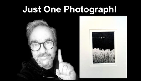 Photographer holding up one framed black and white landscape photograph featuring a dark sky above a textured field.
