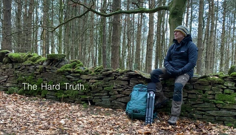 Man sitting on a moss-covered stone wall in a bare forest, with a blue backpack beside him and text overlay reading "The Hard Truth."