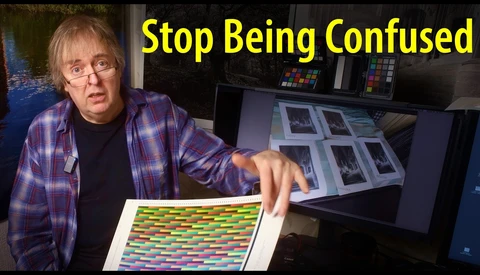Person in plaid shirt gesturing toward color reference charts and monitor displays in a studio setting.