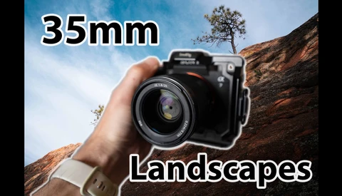 Hand holding a camera with a 35mm lens pointed at a rocky landscape with sparse vegetation.