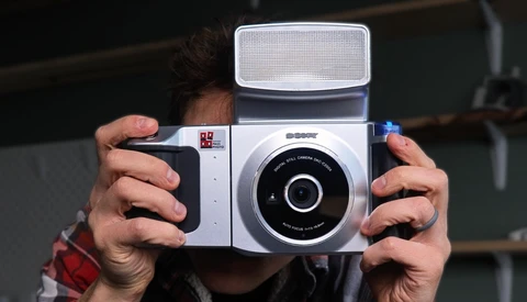 Hands holding a white and black instant film camera with a flip-up flash attachment.