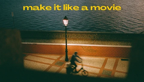 Person sitting on a waterfront boardwalk at dusk beneath a lit street lamp, with calm water and a brick seawall behind them.