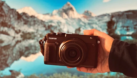 Hand holding a Fujifilm digital camera with snow-capped mountains and a turquoise lake in the background.