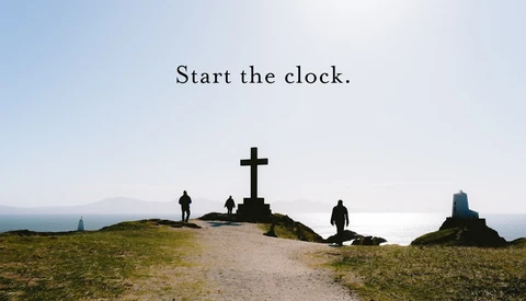 Silhouetted figures stand on a hillside near a large cross overlooking a coastal landscape at dusk.