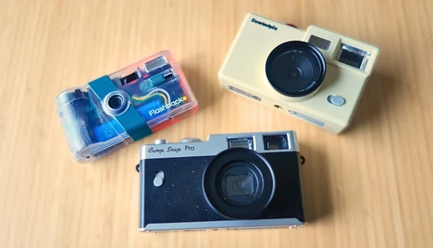 Three instant and film cameras arranged on a wooden surface: a blue disposable camera, a cream-colored instant camera, and a black rangefinder camera.