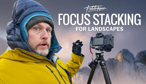 Photographer in winter gear pointing at camera on tripod with misty mountains in background.