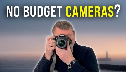 Photographer holding a DSLR camera against a coastal sunset backdrop with a lighthouse visible in the distance.