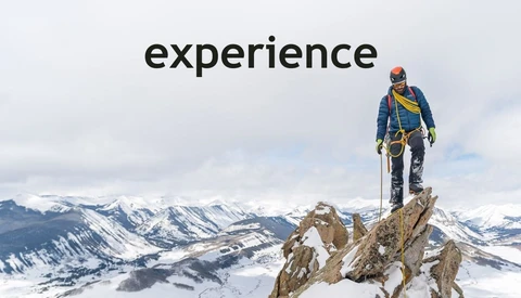 Mountain climber standing on rocky summit overlooking snow-covered alpine landscape.