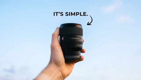Hand holding a black telephoto lens against a blue sky background.