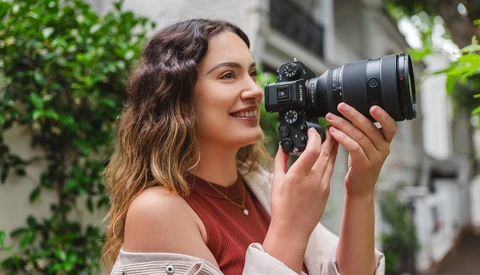 Woman holding a mirrorless camera with telephoto lens, smiling while composing a shot outdoors.