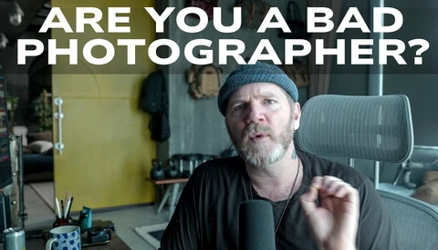 Man with beard and beanie sitting at desk, gesturing while speaking to camera in office setting.