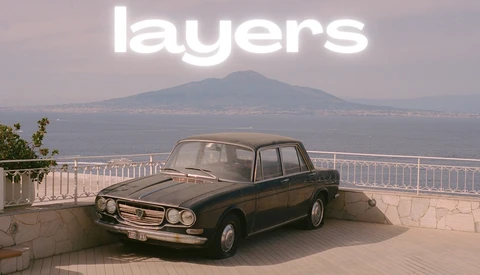 Classic sedan parked on a coastal overlook with a volcanic mountain visible across the water in soft, hazy light.