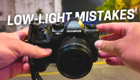 Hands holding an Olympus OM-1 camera with lens against blurred background.