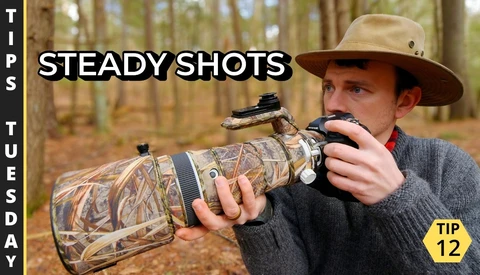 Photographer holding a telephoto lens with camouflage covering while aiming a camera with mounted scope in a forest setting.